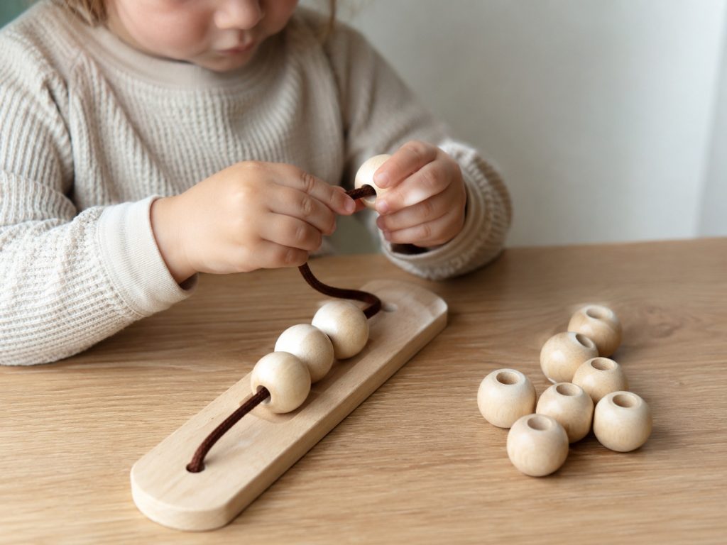 A preschool child threading wooden beads onto a lace during an independent fine motor activity, using classroom toys that build hand-eye coordination and concentration.